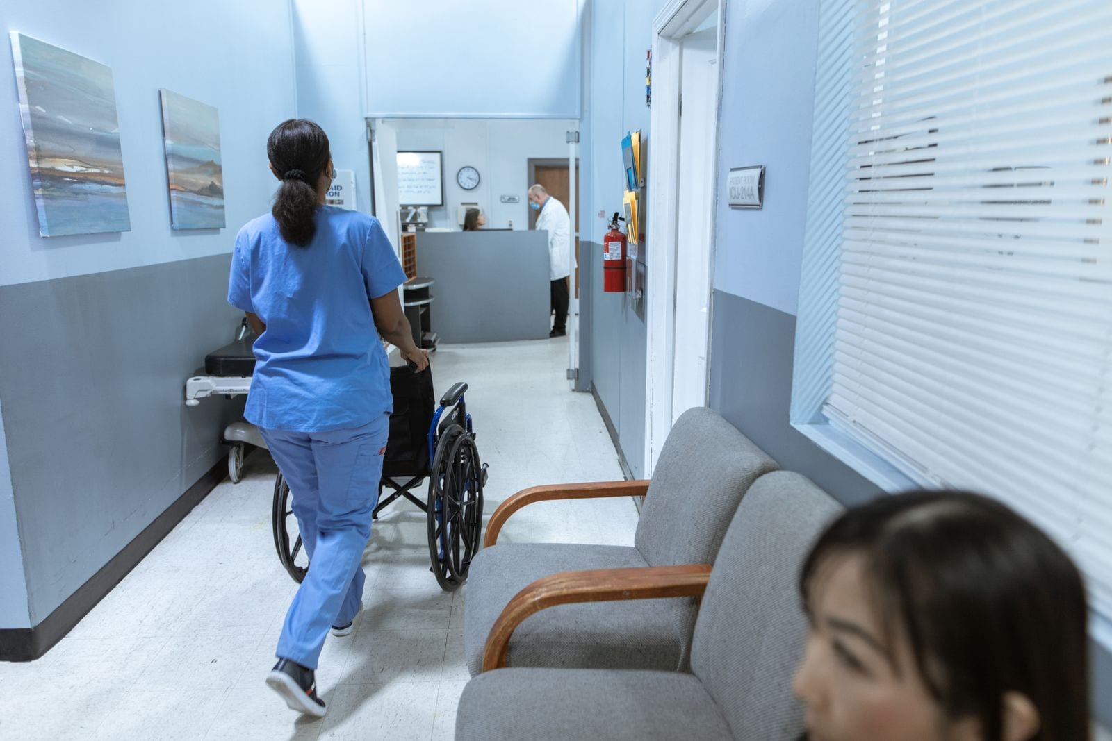 A nurse in scrubs moving a wheelchair along a clinic corridor past a reception desk