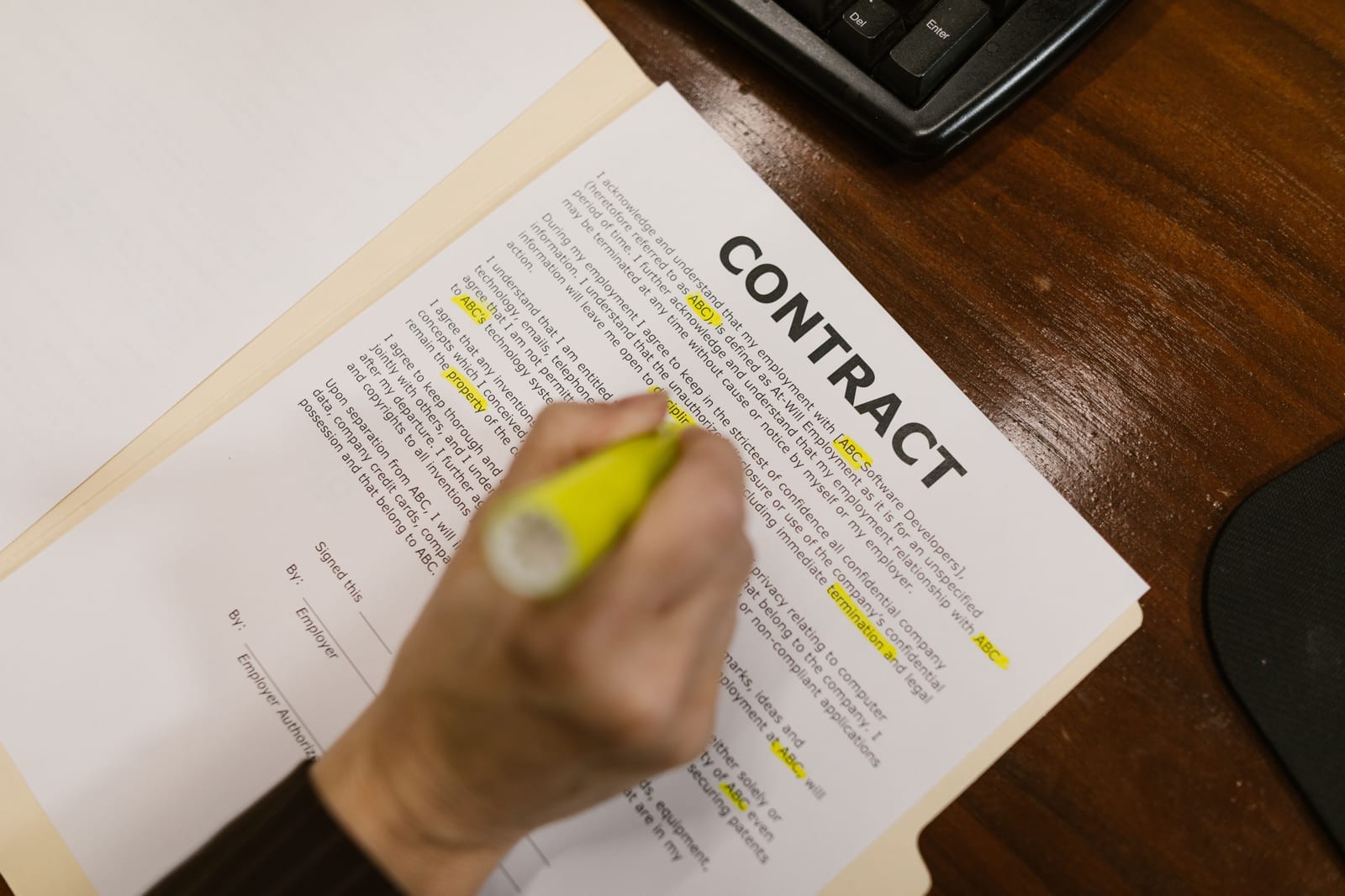 A hand marking a page of contract fine print with a yellow highlighter, in a manila folder on a desk